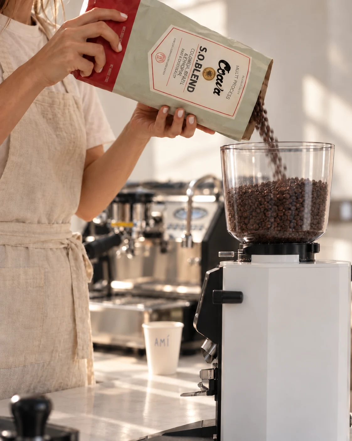 Side view of a white Anfim grinder as a barista pours Coava coffee from a tilted bag, with a La Marzocco GS3 behind